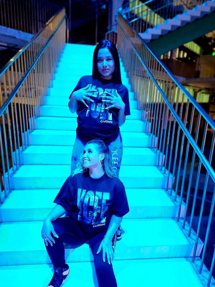 Two people wearing black t-shirts with visible text sitting on a staircase under blue lighting.