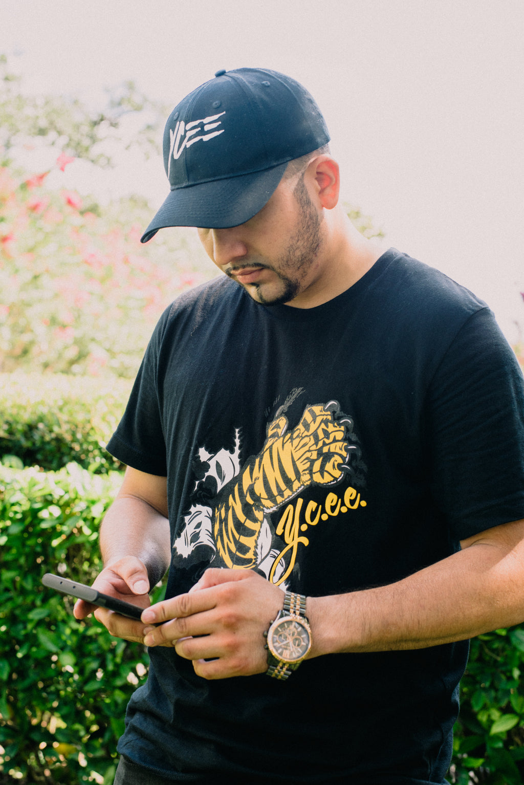 Man wearing a black cap and black t-shirt with a paw design, using a phone outdoors.