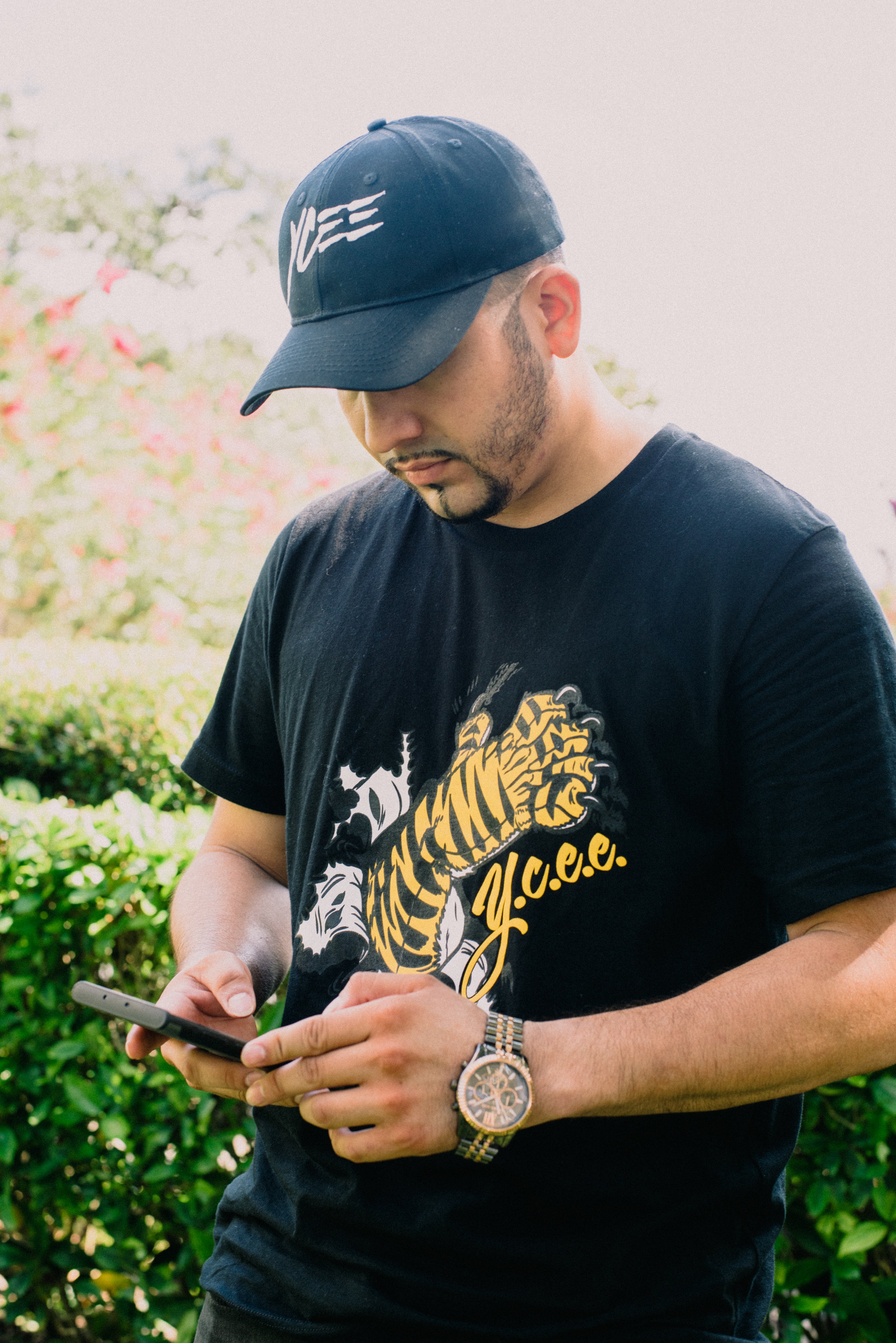 Man wearing a black cap and black t-shirt with a paw design, using a phone outdoors.