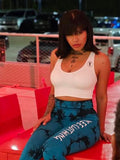 A woman wearing a white crop top and blue patterned leggings sits on a red outdoor chair at night, with a parking lot and cars visible in the background. The foreground shows the back of another person holding a phone.